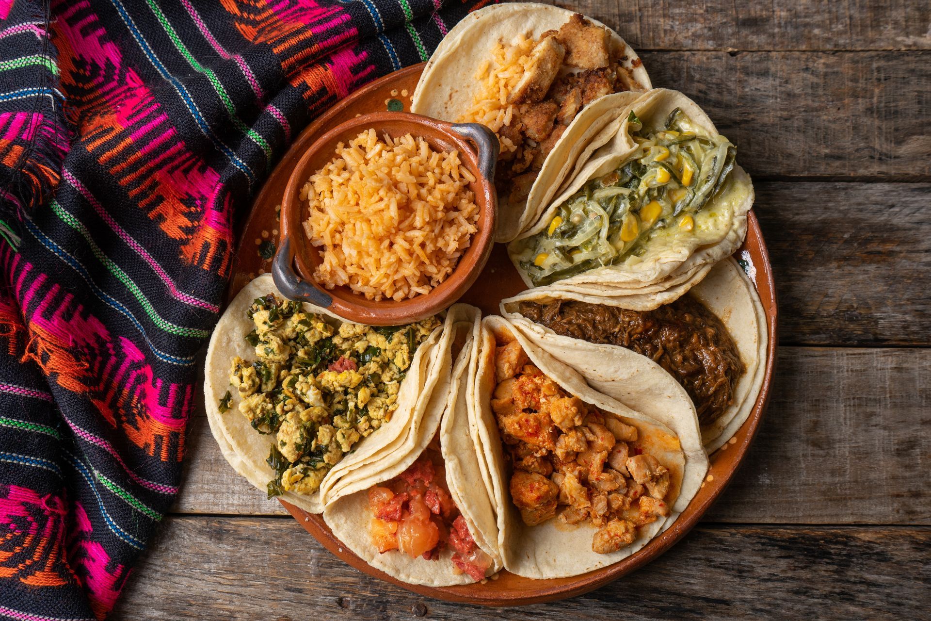 Assorted tacos and side dishes on a wooden table beside a colorful woven cloth.
