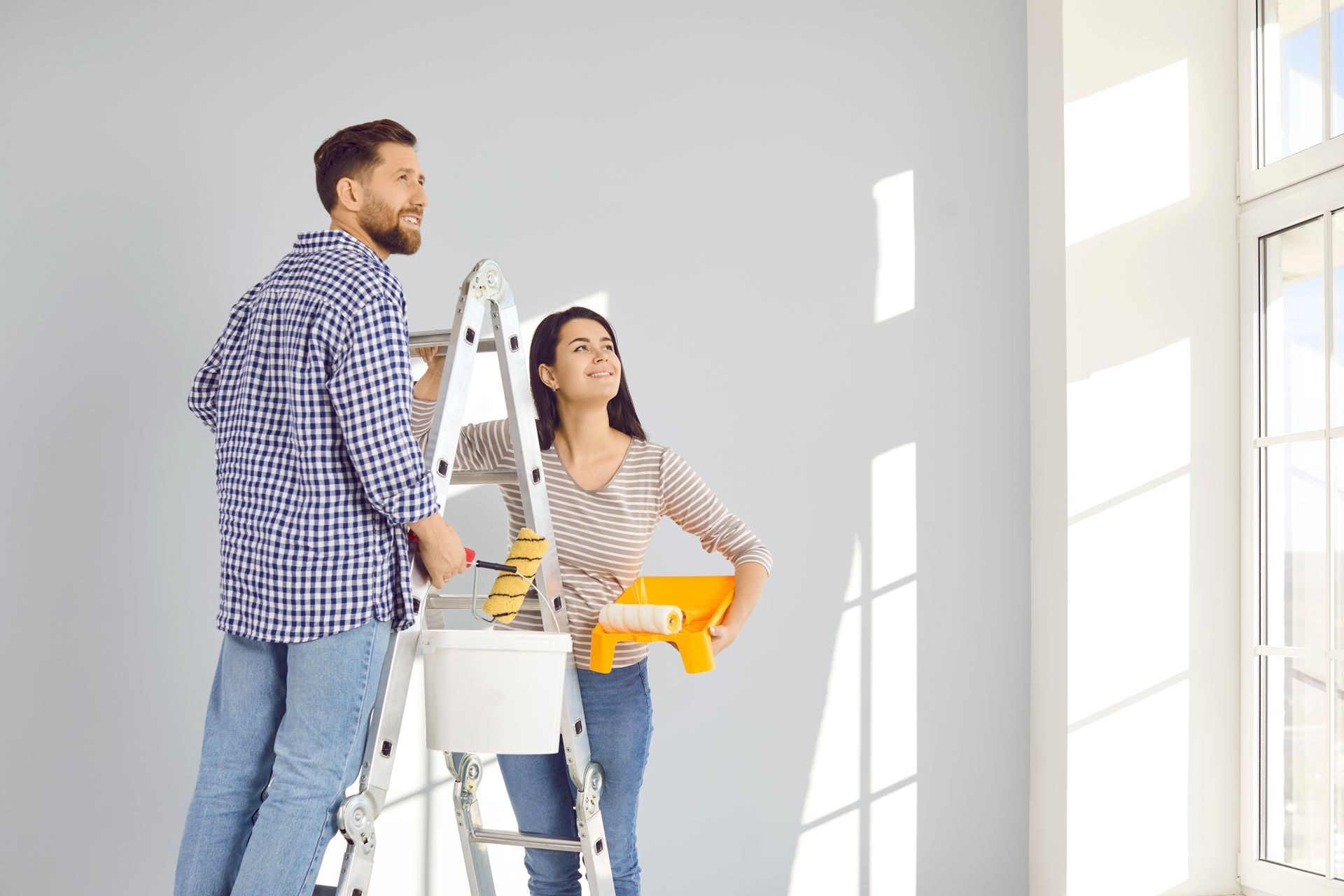A man and a woman are standing on a ladder painting a wall.