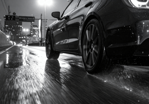 Black car driving on a wet city road at night, water spraying from tires with reflections on the pavement.