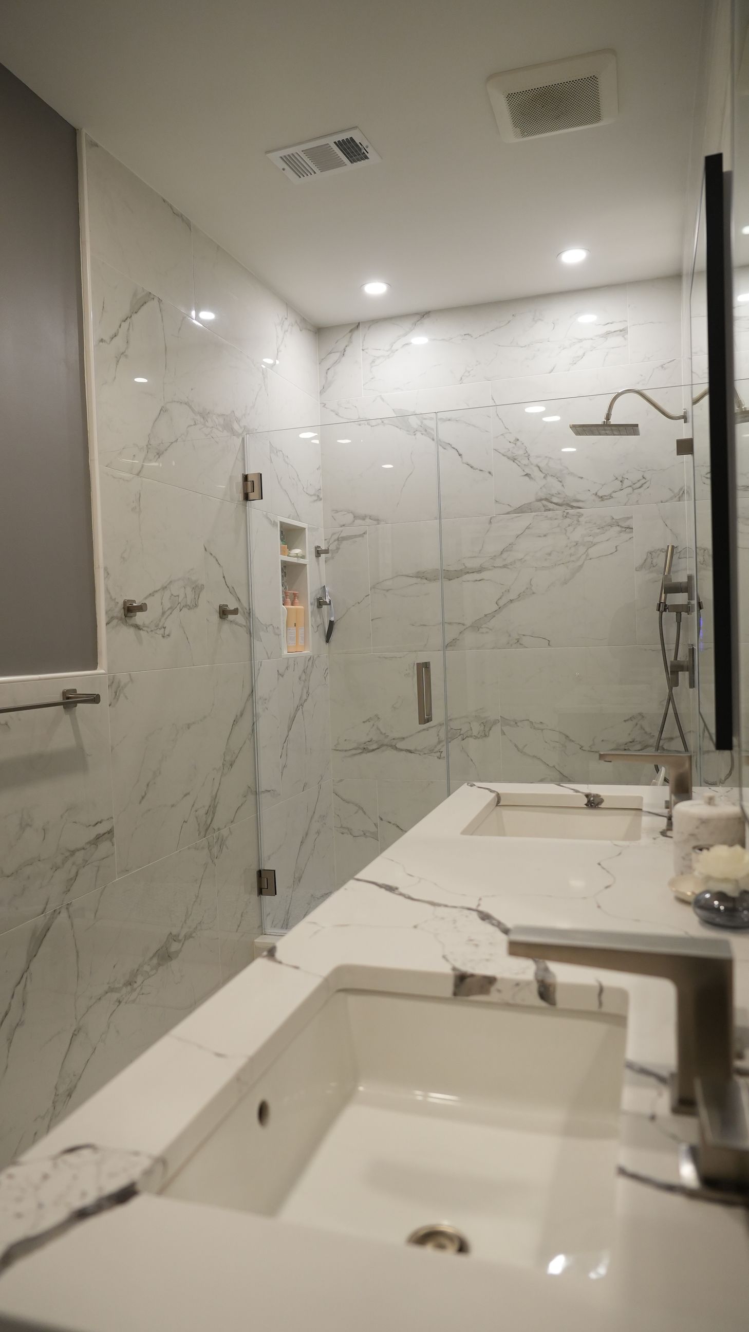 Modern bathroom with marble walls and a glass shower. Two sinks sit on a white countertop.
