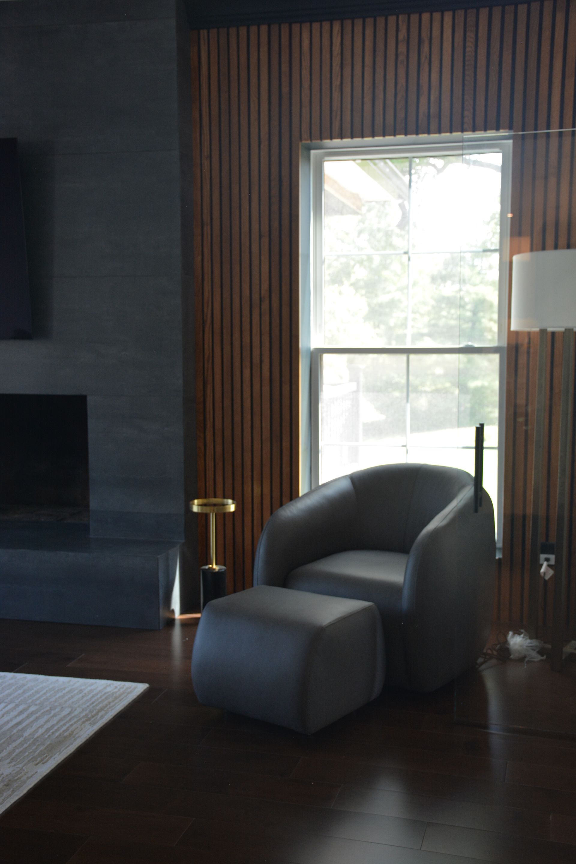 Gray armchair and ottoman in a room with wood paneling, window, and a black fireplace.