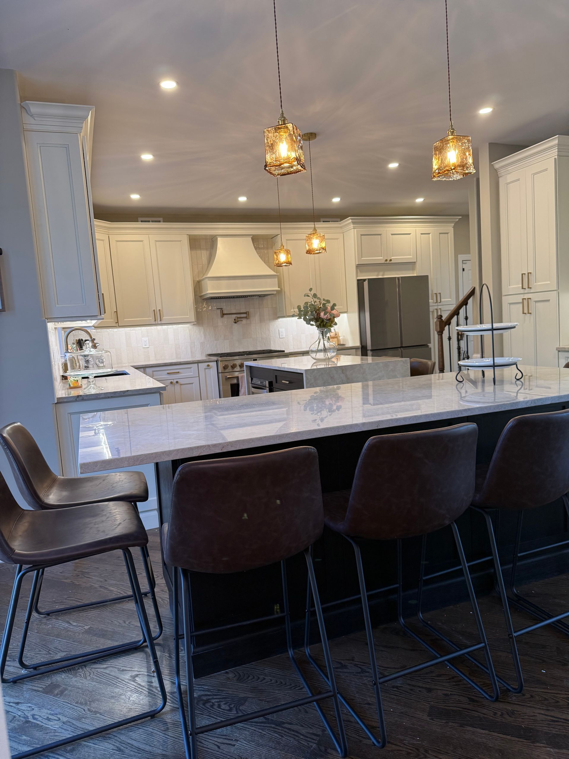 A modern kitchen with a large island and pendant lights. Brown leather bar stools. Light gray cabinets and countertops.