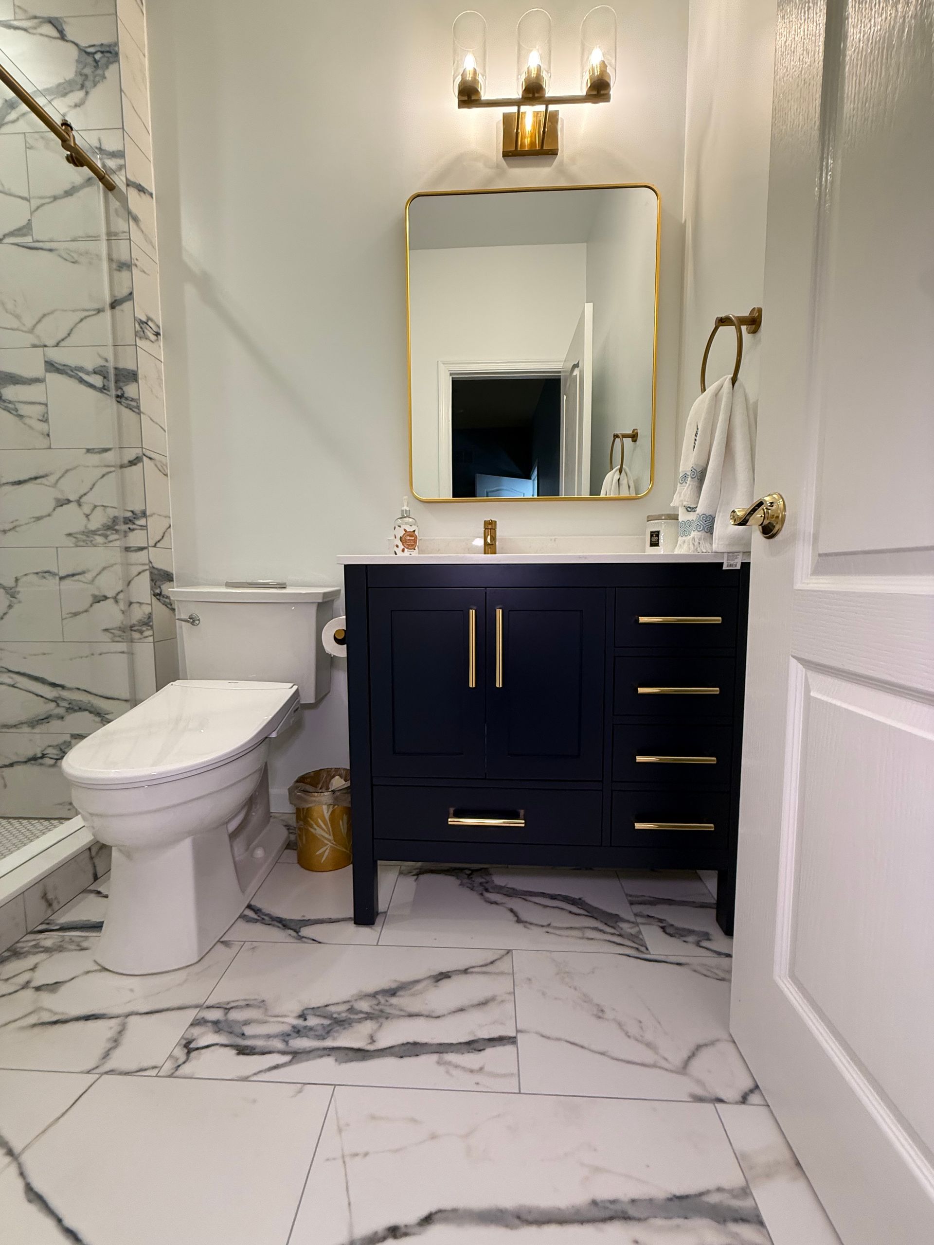 Bathroom with marble flooring and shower, navy vanity with gold hardware, and a gold-framed mirror.