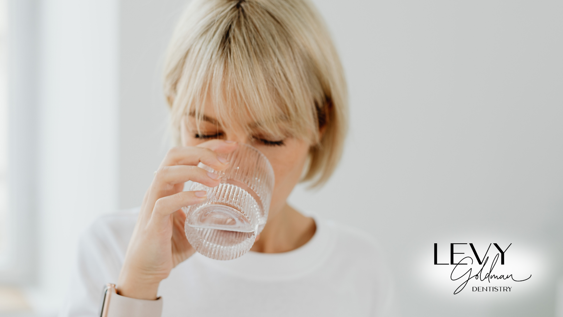 Blonde woman drinking water from a clear glass, eyes closed, white background.