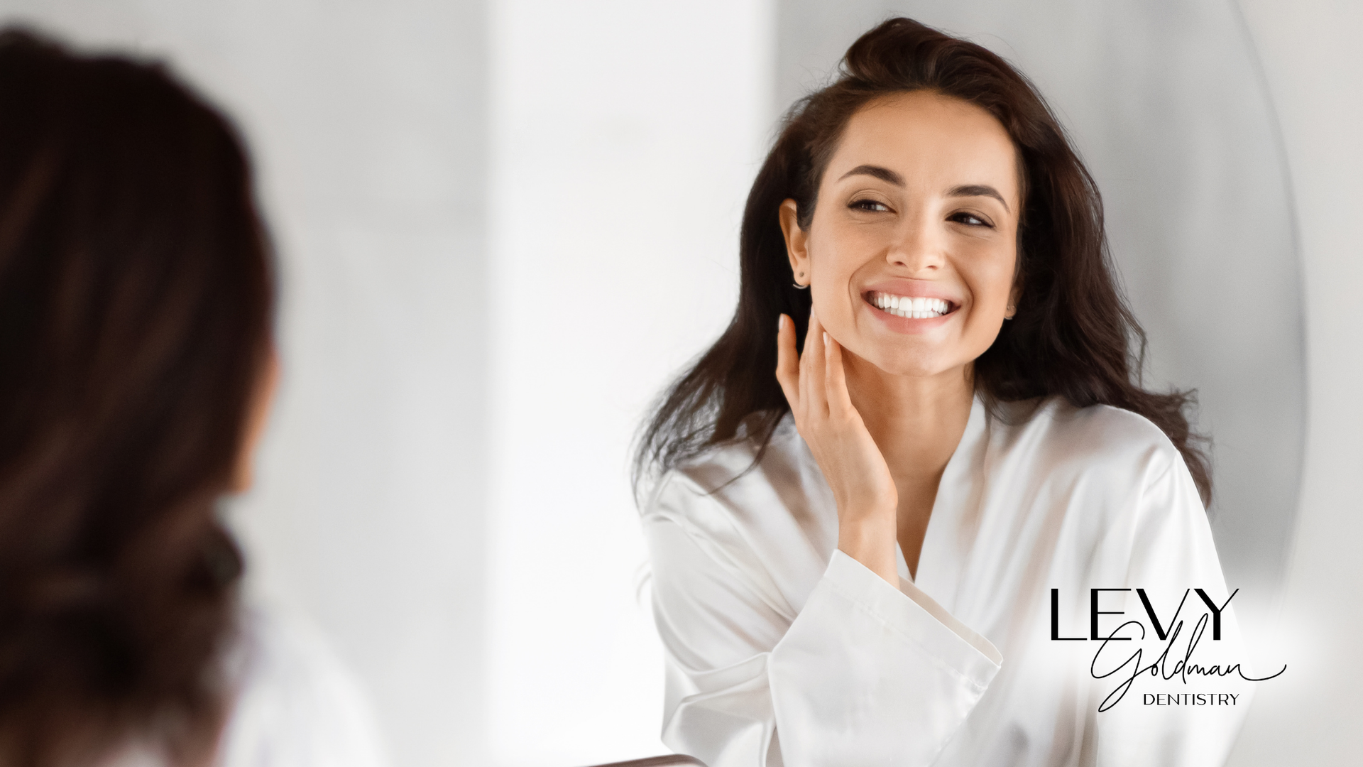 Woman in white robe smiling in front of a mirror, touching her face.