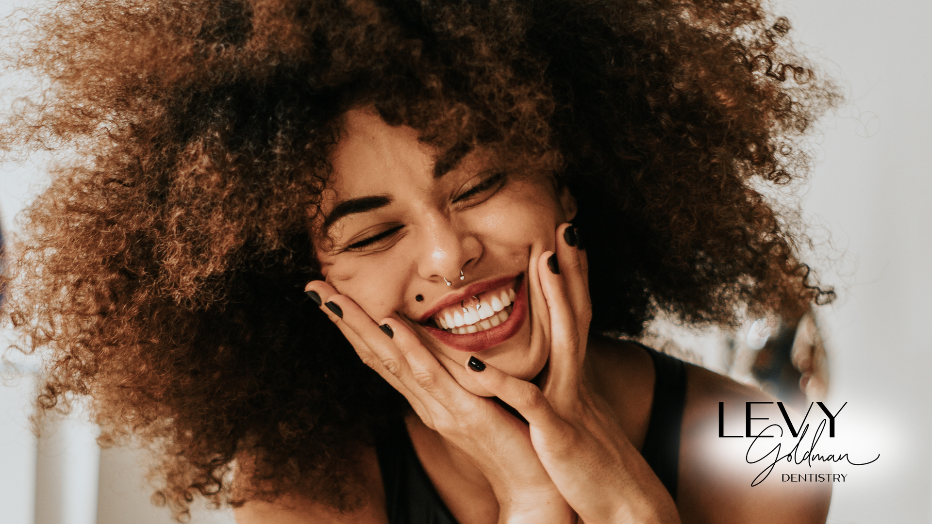 A woman with curly hair is smiling with her hands on her face.