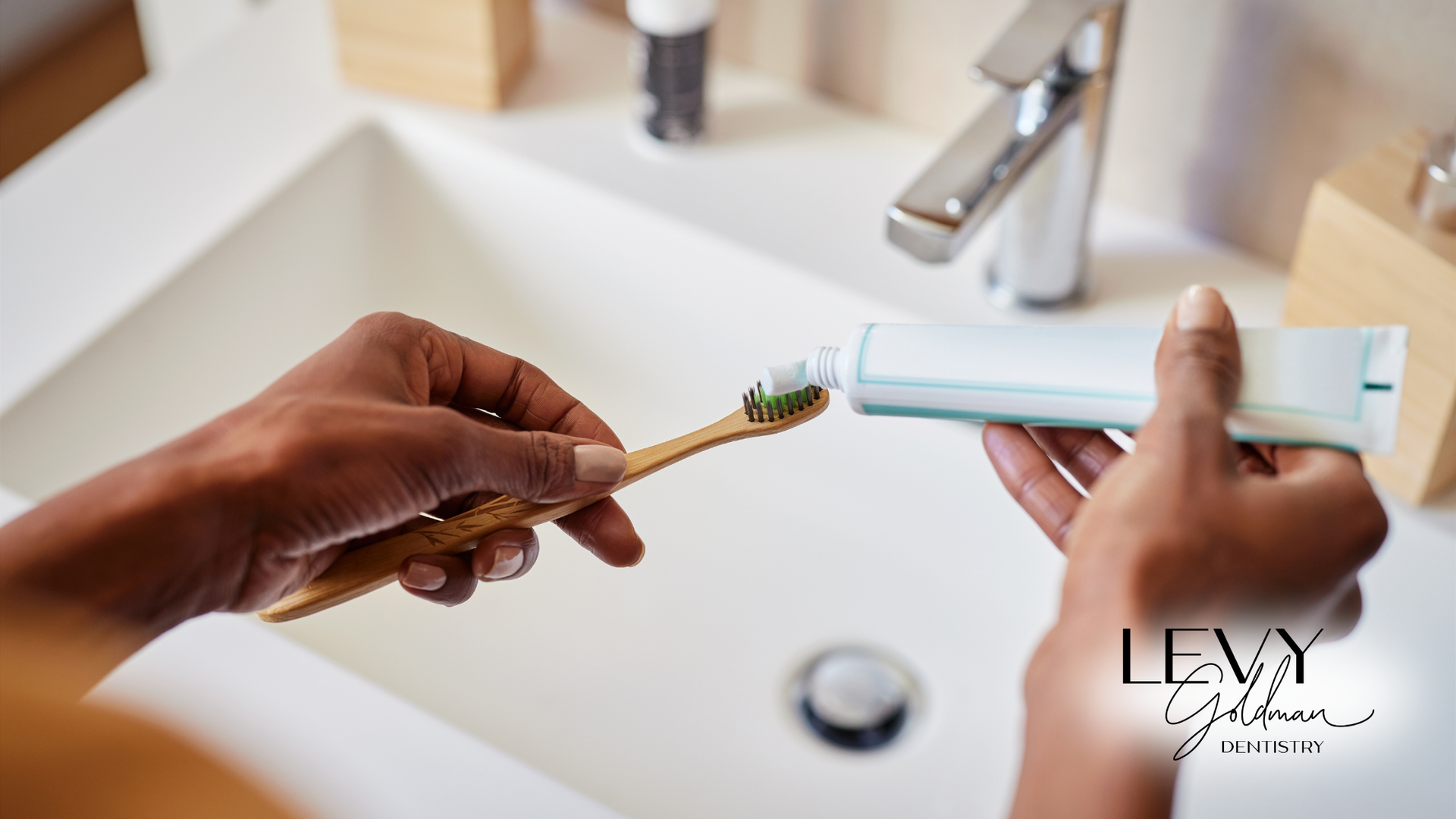 A person is putting toothpaste on a wooden toothbrush in a bathroom sink.