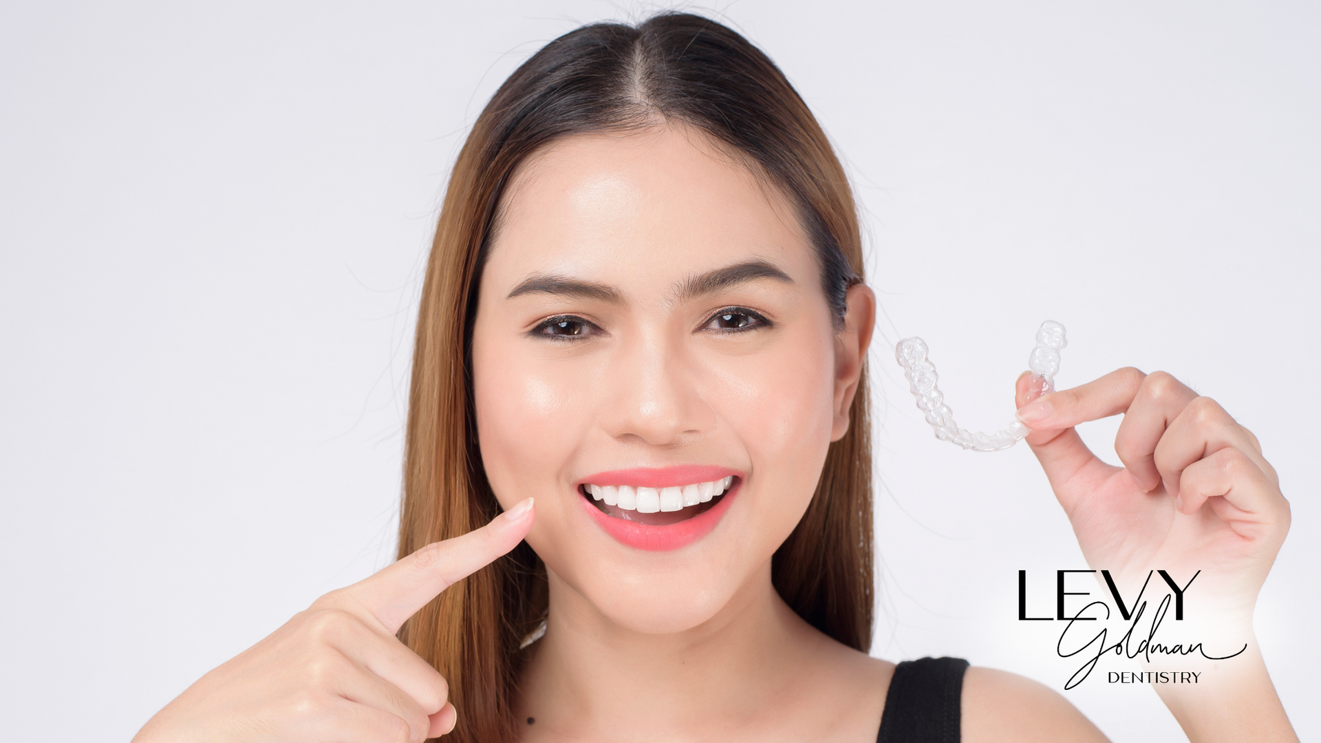 A woman is smiling and pointing at her teeth while holding Invisalign.