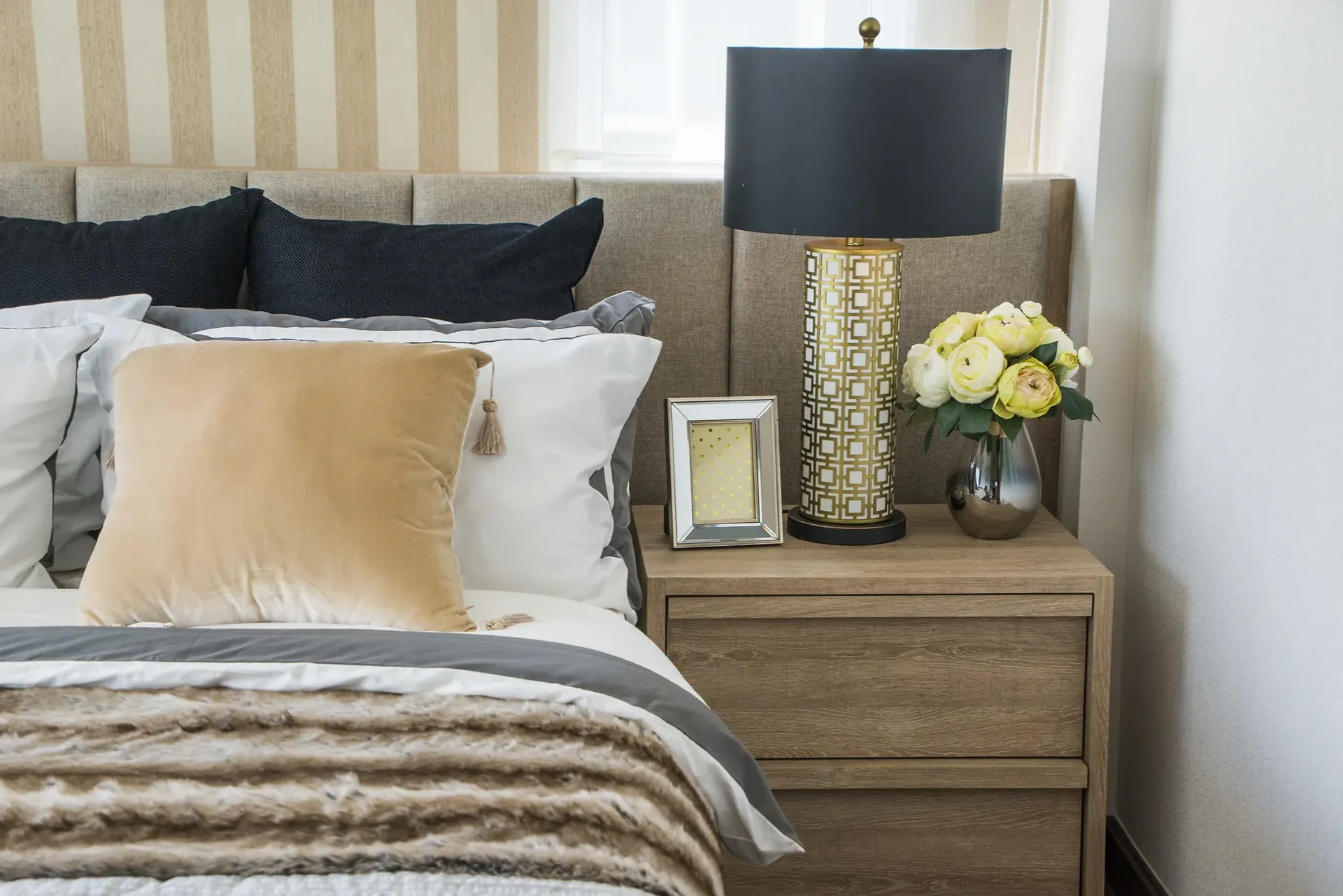 A bedroom with a bed , nightstand , lamp and vase of flowers.