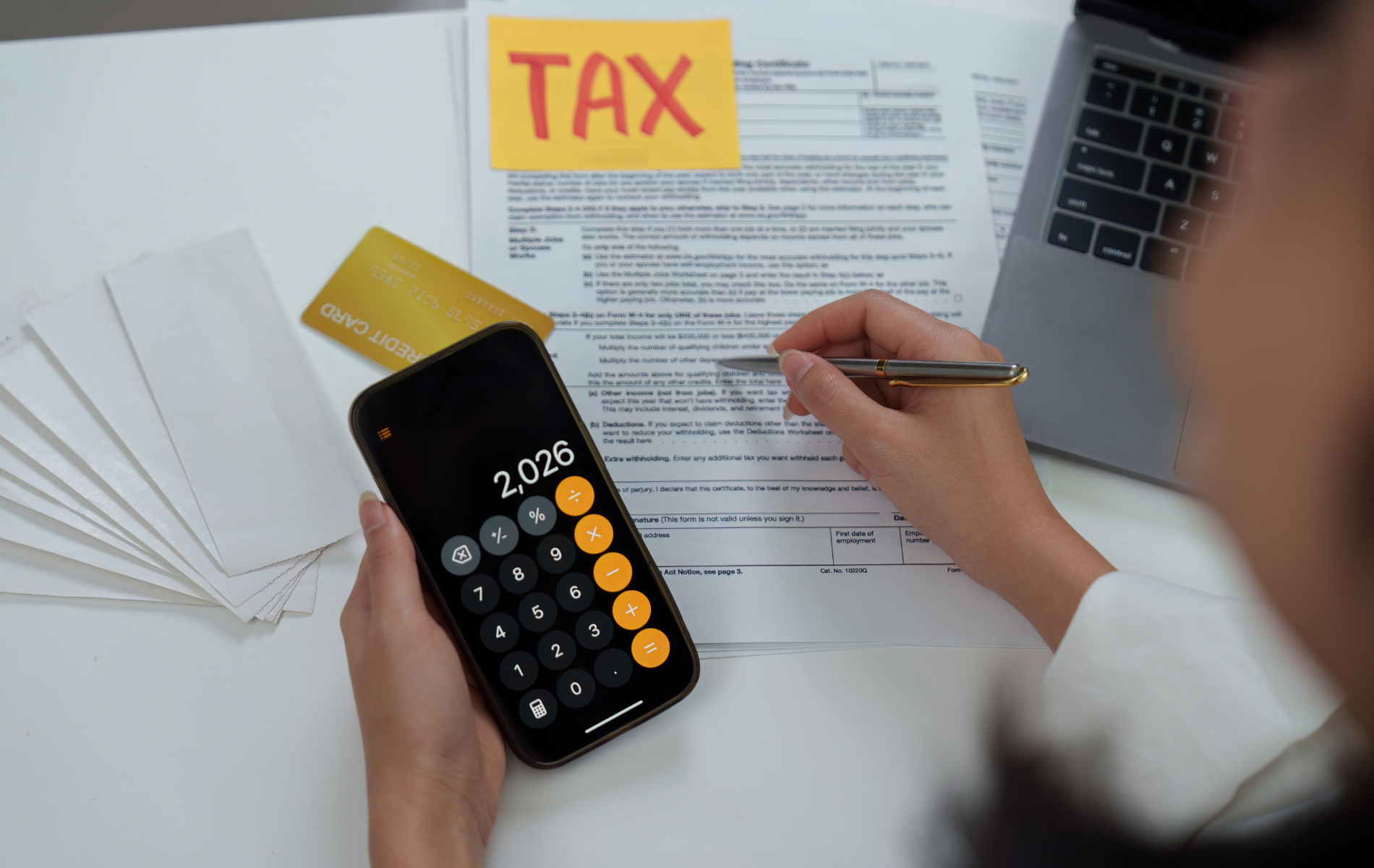 Person using a calculator and pen to fill out tax forms, with a credit card, laptop, and envelopes visible.