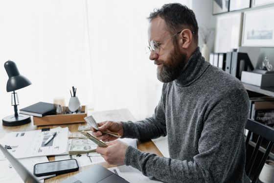 Man counting money at a desk with papers, lamp, and laptop.