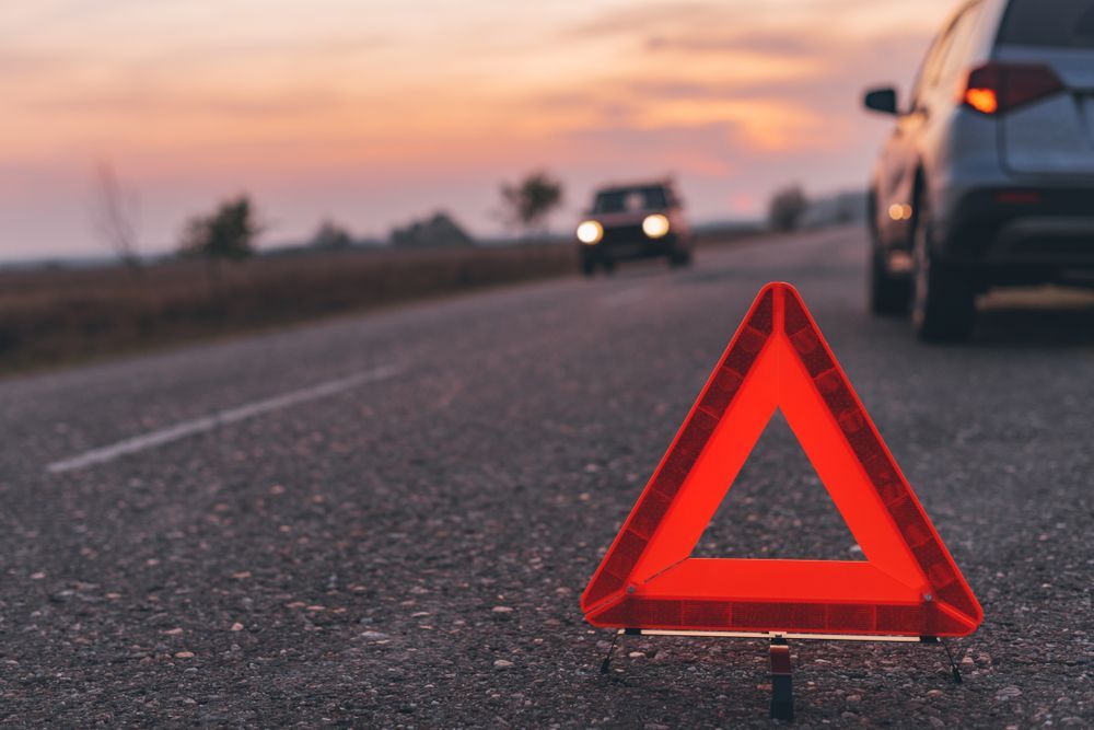 Red warning triangle on a dark asphalt road, with a car stopped and another approaching in the distance at sunset.