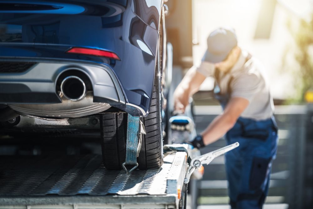 A tow truck operator secures a blue car on a flatbed, using straps. The scene takes place outdoors.