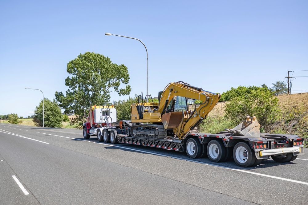 A red semi-truck hauling a yellow excavator on a flatbed trailer, driving on a sunny road.
