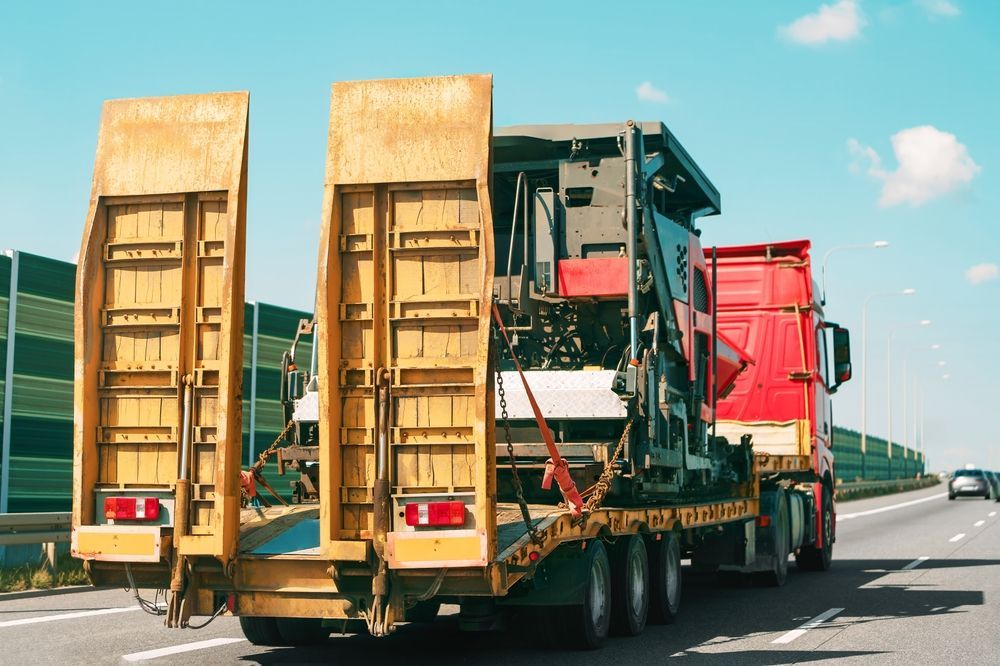 A large, yellow and red flatbed truck hauling machinery on a highway with green sound barriers.
