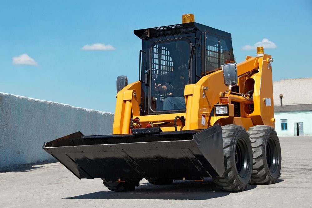 Yellow skid steer loader with black bucket, parked on pavement against a blue sky.