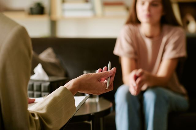 Counselor holding a pen across from a blurred seated client in a calm office setting Therapist gesturing with a pen while speaking to a seated client in a 상담 room