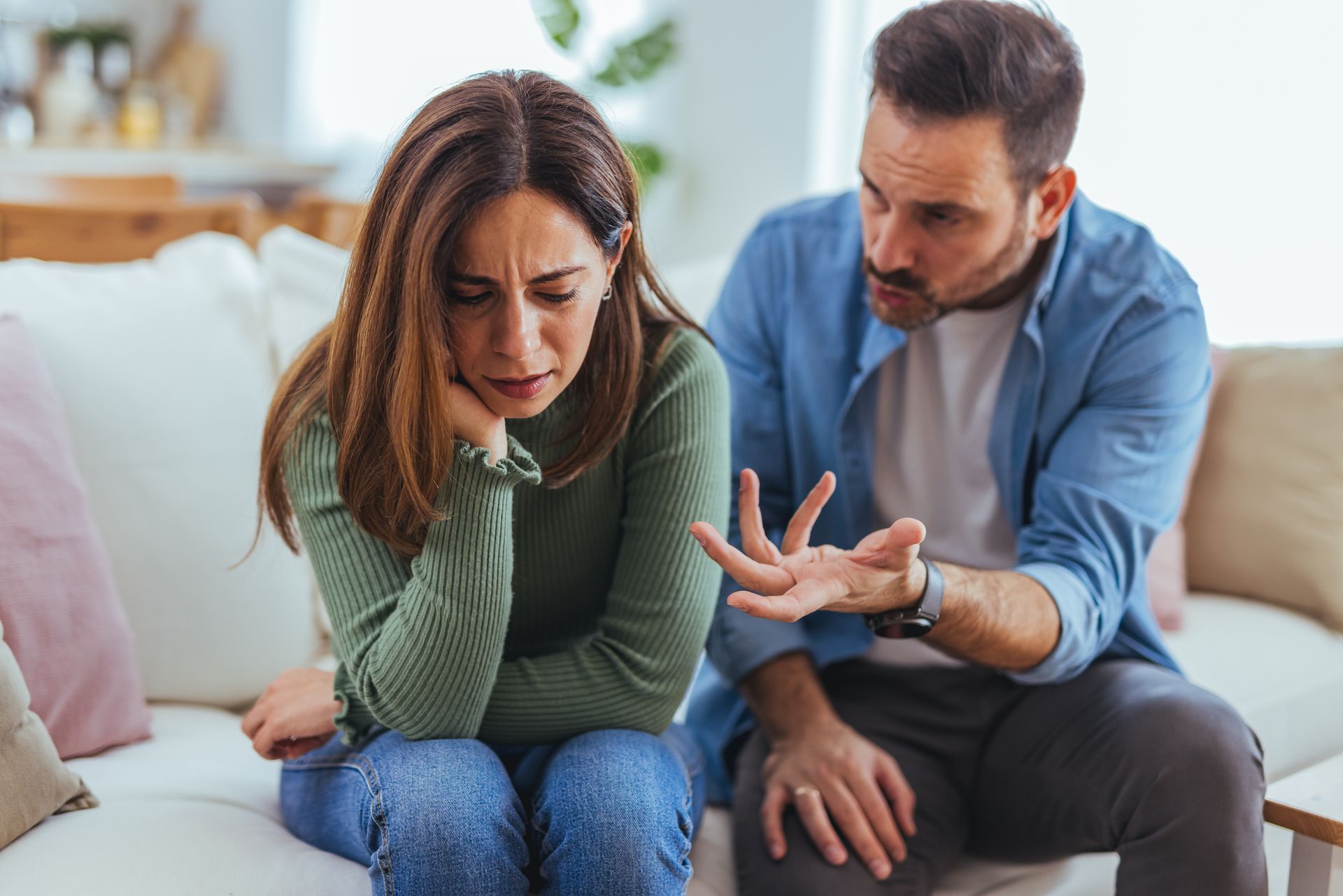Two people talking on a couch, one gesturing with a raised finger in a bright living room Crying woman on couch while man beside her gestures during a tense conversation