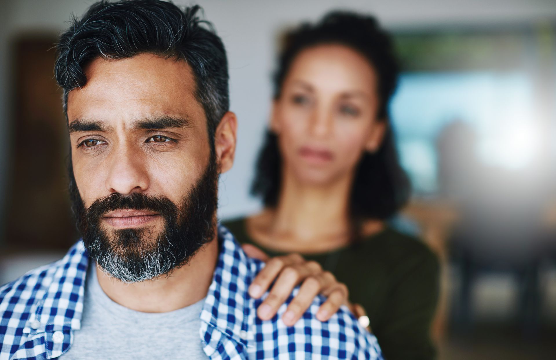 Therapist talking with a client on a couch in a bright living room office A bearded man in a checkered shirt looks down while a woman stands behind him with a hand on his shoulder.