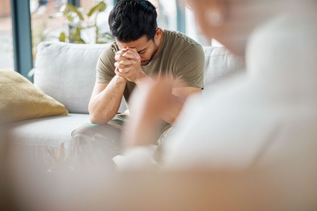 Person sitting on a couch with head bowed and hands clasped, looking distressed in a softly lit room Man sitting on a couch with head bowed and hands clasped, looking distressed in a bright living room