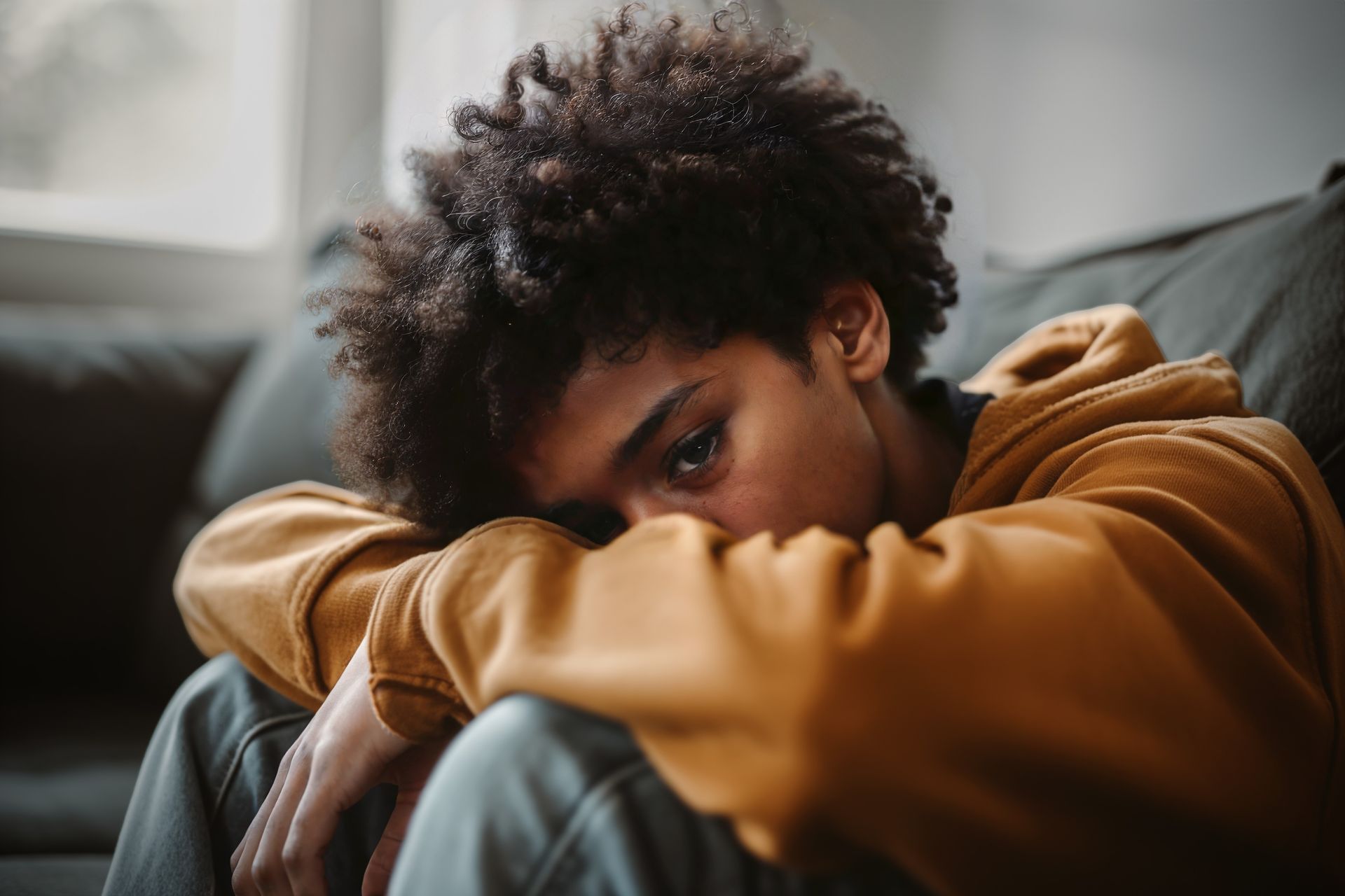 Person in a mustard hoodie resting their head on folded arms on a couch, looking down sadly.