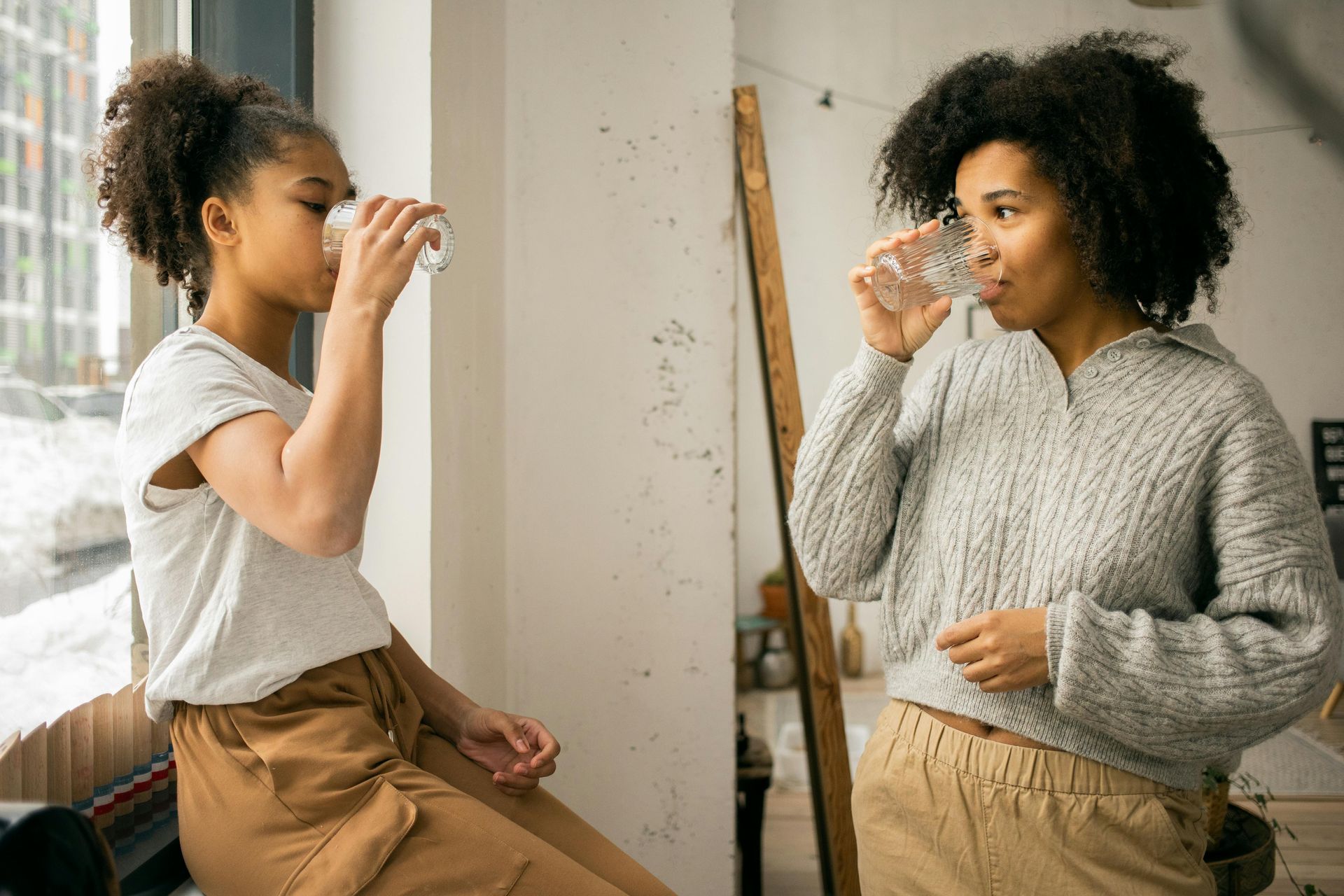 Two women are drinking water from glasses in front of a window.