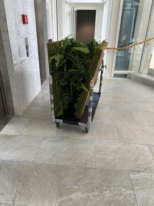Un chariot à bagages dans un hall lumineux transportant des panneaux recouverts de feuillage artificiel vert.