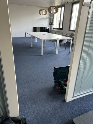 Un espace de bureau avec une grande table blanche, une moquette bleue, deux lampes suspendues et une boîte à outils posée au sol près de la porte.