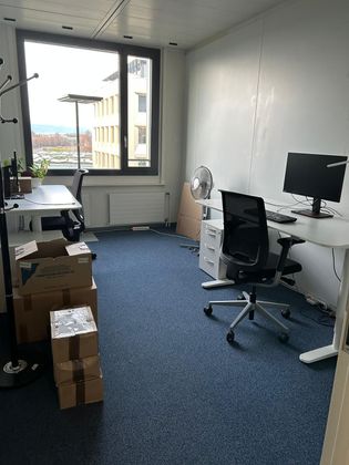 Un petit bureau bien rangé avec deux bureaux, des chaises ergonomiques, une grande fenêtre et des cartons empilés sur une moquette bleue.