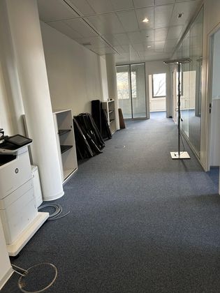 Un couloir de bureau vide avec de la moquette grise, une imprimante blanche au premier plan, des étagères de rangement et des cloisons vitrées.