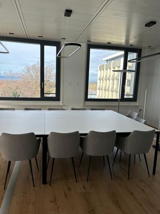 Une salle de conférence moderne avec une grande table rectangulaire blanche entourée de chaises grises et des fenêtres offrant une vue agréable.