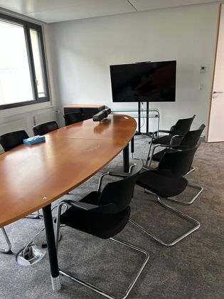 Dans un bureau, une table de conférence en bois et des chaises noires sont disposées, avec un grand écran sur un support contre le mur.