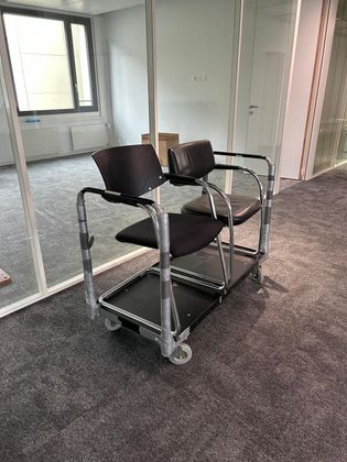 Deux chaises noires fixées sur un chariot de transport métallique à roulettes dans un couloir de bureau.