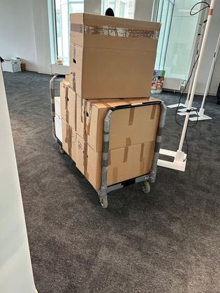 Dans un bureau à la moquette grise, une pile de cartons de déménagement bruns repose sur un chariot métallique.