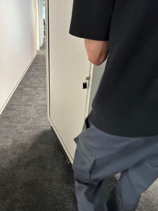 Une personne vêtue d'une chemise noire et d'un pantalon cargo gris se tient près d'une porte dans un couloir recouvert de moquette.