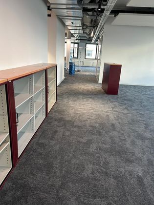 Un espace de bureau moderne avec moquette grise, conduits d'aération apparents au plafond, murs blancs, étagères de rangement et une armoire bordeaux.