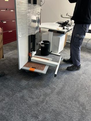 Une personne en tenue de travail se tient à côté d'un présentoir blanc à roulettes en cours d'assemblage ou de déplacement dans un bureau.