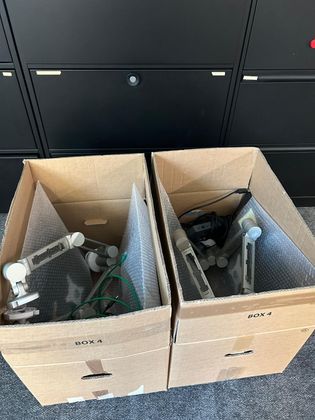 Deux boîtes en carton ouvertes posées sur un sol recouvert de moquette, contenant chacune une lampe de bureau blanche réglable, emballée dans du papier bulle.