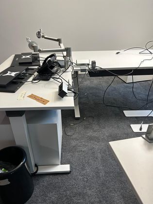 Un espace de travail composé d'un bureau blanc, d'un bras pour écran, d'appareils électroniques et de câbles emmêlés, le tout sur un sol recouvert de moquette grise.