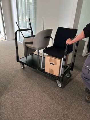 Une personne pousse un chariot utilitaire noir contenant deux chaises — une beige, une noire — et une boîte en carton dans un espace de bureau.