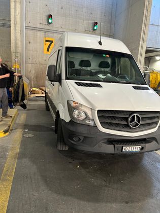 Une camionnette Mercedes-Benz Sprinter blanche garée dans un garage intérieur à côté d'une personne se tenant près d'un mur portant le numéro 7.