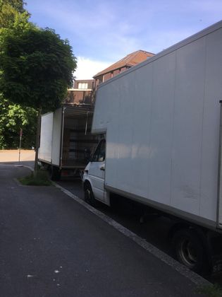 Deux gros camions blancs étaient garés l'un derrière l'autre le long d'un trottoir, à côté d'un arbre vert et d'un immeuble résidentiel.