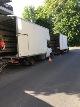 Deux camions de déménagement blancs garés en file indienne dans une rue ensoleillée, portes arrière ouvertes, et des cônes de signalisation orange à proximité.