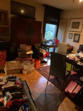 Un bureau à domicile encombré, avec une armoire en bois sombre, un bureau recouvert de papiers, une chaise et plusieurs cartons au sol.