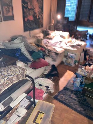 Un salon encombré avec un canapé recouvert de coussins, de couvertures, de vêtements et d'objets divers, ainsi que des affaires éparpillées.