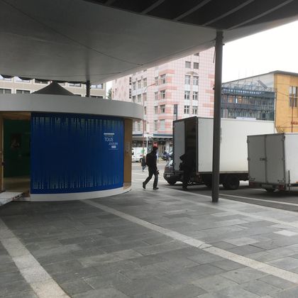 Une personne passe devant un pavillon circulaire bleu incurvé, situé sur une place pavée, à côté de camions stationnés et de bâtiments municipaux.