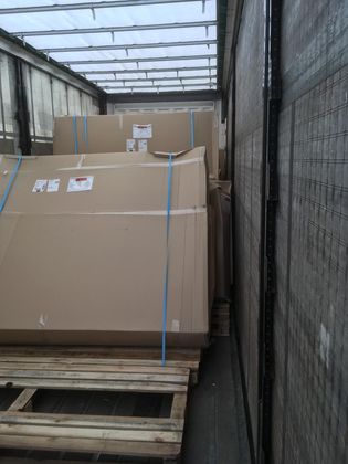 De grandes caisses en carton, fixées par des sangles bleues sur des palettes en bois, se trouvent à l'intérieur d'une semi-remorque.