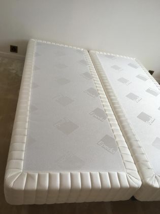 Deux sommiers blancs identiques, recouverts de tissu, sont placés côte à côte sur un sol moquetté, contre un mur uni.