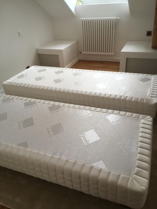 Deux ensembles de matelas jumeaux identiques placés côte à côte dans une chambre aux murs blancs, avec un radiateur et des tables de chevet intégrées.