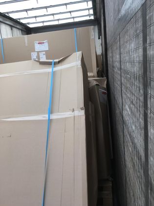 De grandes piles de matériaux emballés dans du carton sont chargées sur la benne d'un camion et sécurisées par des sangles bleues.